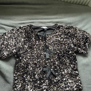 Svart paljettopp  - Glittrig svart topp från Gina Young, täckt med silvriga paljetter och dekorerad med tre svarta rosetter framtill. Kortärmad modell med rund hals och croppad passform. Tröjan är endast använd en gång så gott som ny. 