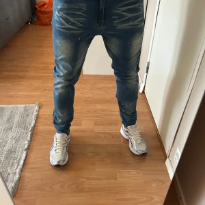 NVLTY Jeand - Säljer ett par blå jeans från NVLTY i storlek 34. Jeansen har coola slitningar och blekta partier framtill och baktill, samt små vita färgstänk för en trendig look. Tillverkade i stretchig bomullsmix för skön passform.