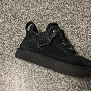 Svarta sneakers från UGG i mocka och mesh - Snygga helsvarta sneakers från UGG med ovandel i mocka och mesh, breda snören och rund tå. Skorna har en platt sula och dragflik bak för enkel påtagning. Perfekta för dig som gillar en clean och stilren look.