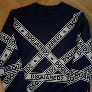 Blå DSQUARED2 sweatshirt med logga - Snygg marinblå sweatshirt från DSQUARED2 med stora vita loggor diagonalt över hela tröjan. Tröjan har rund hals och långa ärmar. Perfekt för dig som gillar streetwear och vill sticka ut med en trendig look.