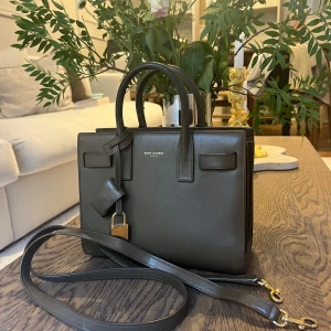 Saint Laurent Sac de Jour handväska - Snygg och stilren handväska från Saint Laurent i en khaki grön färg.  Klassisk boxig form med dubbla handtag, avtagbar axelrem och guldfärgat lås. Det finns lite slitage, som ni kan se på bilden. Inget man riktigt tänker på om man inte kollar nära. Väskan tål väldigt mycket. Väskan är i en mindre storlek, Saint Laurent Sac de jour nano. Nypris 23 000 kr. Hör mer än gärna av er för fler frågor!😊