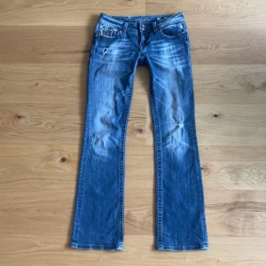 Miss Me bootcut jeans - Snygga blå jeans från Miss Me med bootcut passform. Jeansen har slitna detaljer, dubbla knappar och coola bakfickor med spets och bling. Perfekta för dig som gillar statement-jeans med unika detaljer.