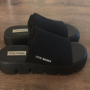 Svarta chunky sandaler Steve Madden - Chunky svarta sandaler från Steve Madden med bred ribbad rem över foten och kraftig plattformsula. Snyggt minimalistisk design med öppen tå och logga på sidan. Perfekta för en trendig look under varma dagar.