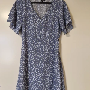 Blå blommig kortklänning med volangärm - Supersöt blå kortklänning med vitt blommönster, v-ringning och knappar framtill. Klänningen har volangärmar och resår i midjan för en snygg passform. Perfekt för vår och sommar!