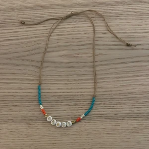 Färgglatt pärlhalsband med texten PEACE - Ett handgjort halsband med beige snöre och färgglada pärlor i blått, vitt, orange och guld. I mitten sitter vita pärlor med guldbokstäver som bildar ordet PEACE. Justerbar längd med knytning för perfekt passform.