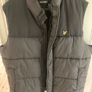 Svart dunväst från Lyle & Scott - Säljer en svart dunväst från Lyle & Scott med klassisk gul logga på bröstet. Västen har hög krage, dragkedja och tryckknappar framtill samt två stora fickor. Perfekt för lager-på-lager och riktigt skön att bära under kyliga dagar.