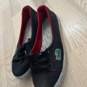 Svarta ballerinaskor från Lacoste - Svarta ballerinaskor från Lacoste med röd kant och snörning framtill. Skorna har klassisk rund tå, vit sula och den ikoniska gröna krokodilloggan på sidan. Insidan är fodrad med rött tyg och innersulan har Lacoste-tryck. Perfekta för en sportig och stilren look.