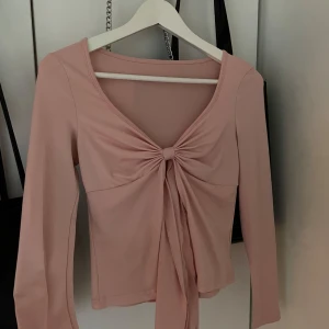 Rosa långärmad topp med knyt - Supersöt ljusrosa långärmad topp med snygg knytning framtill och djup v-ringning. Toppens material är mjukt och stretchigt, perfekt för en trendig look. Passar perfekt till både jeans och kjol för en cool outfit.
