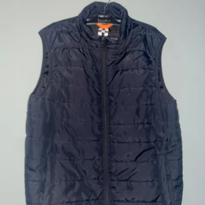 Svart quiltad dunväst från X-Trail - Svart quiltad dunväst från X-Trail i storlek S. Västen har hög krage och dragkedja framtill. Perfekt att slänga över en hoodie eller tröja när det är kyligt ute. Enkel och stilren design som funkar till det mesta. 