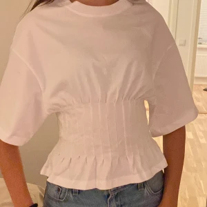 Vit korsett-topp - Säljer en vit topp med korsett-inspirerad midja och korta ärmar. Toppen har rund halsringning och snygga sömdetaljer som ger en markerad siluett. Perfekt att styla med jeans för en trendig look. Endast använd 2 gånger.