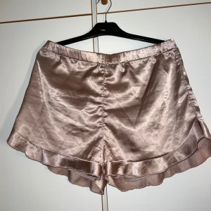 Champagnefärgade satinskjorts Boohoo - Snygga champagnefärgade shorts från Boohoo i glansigt satinmaterial. De har en loose passform, elastisk midja och volangkant nedtill för en extra feminin touch. Perfekta för sommarkvällar eller när du vill känna dig extra lyxig.