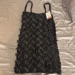 Svart kort klänning med fransar - Supersnygg svart kort klänning med breda axelband och massor av fransdetaljer över hela klänningen. Perfekt för dig som vill sticka ut med en cool och trendig look. Klänningen har en rak passform och är riktigt festlig.