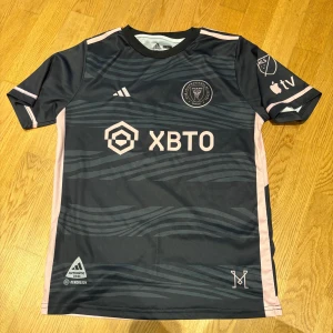 Inter Miami Messi matchtröja Adidas - Snygg Inter Miami CF fotbollströja från Adidas med Messi och nummer 10 på ryggen. Tröjan är svart med ljusrosa detaljer och vågiga mönster på framsidan. Korta ärmar, rund hals och tillverkad i lätt funktionsmaterial. Perfekt för dig som älskar fotboll och Messi.