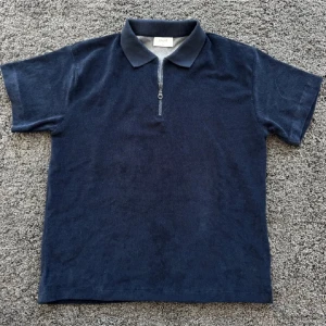 Mörkblå pikétröja från grunt - Snygg mörkblå pikétröja från grunt med klassisk krage och half zip-dragkedja framtill. Tröjan har korta ärmar och är tillverkad i ett mjukt material som känns skönt mot huden. Perfekt för en stilren och avslappnad look. Aldrig använd 