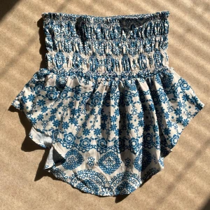 Blåvit bandeau tubtopp med mönster - Bandeau tubtopp i blått och vitt med mönster och smockad överdel. Toppen har volangkant nedtill och är perfekt för varma  sommar dagar. Känns lätt och luftigt.