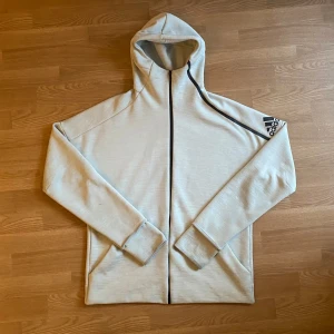 Adidas Z.N.E hoodie i färgen ljusgrå/benvit - Snygg ljusgrå/benvit hoodiejacka från Adidas med svart logga på ärmen och svarta detaljer längs dragkedjan samt 3m detaljer. Tröjan är lite översized i storlek, har huva, långa ärmar och två fickor framtill. Perfekt för chill dagar eller när du vill ha en sportig look. Köptes för 1200kr!