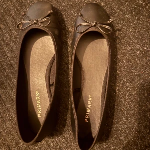 Svarta ballerinaskor från Primark - Klassiska svarta ballerinaskor från Primark med rund tå och söt rosettdetalj framtill. Skorna har platt sula och är tillverkade i syntetmaterial, perfekta för en stilren och enkel look. Helt nya.