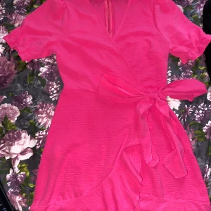 Rosa omlottklänning med volang - Superfin rosa kortklänning med omlottmodell och knyt i midjan. Klänningen har korta puffärmar och volangdetalj längs nederkanten. Perfekt för varma dagar och ger en färgstark look. Materialet känns lätt och luftigt. Använd endast 2 gånger.!