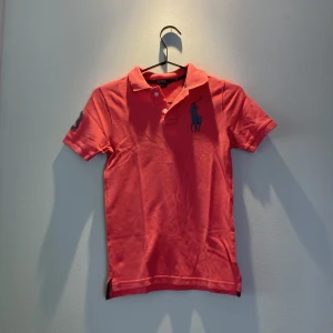 Korallröd pikétröja från Ralph Lauren - Snygg korallröd pikétröja från Ralph Lauren med klassisk krage, knappar och stor marinblå logga på bröstet. På ena ärmen finns en grå broderad siffra 3. Tröjan är i bomull och har korta ärmar, perfekt för en sportig och stilren look.