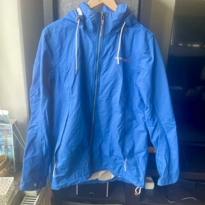 Blå vindjacka från Didriksons - Snygg blå vindjacka från Didriksons med huva, vita snören och dragkedja framtill. Jackan har broderad logga på bröstet och tryckknappar med märkets namn. Perfekt för blåsiga dagar och har en sportig vibe.