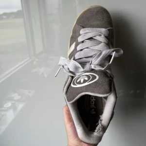 Grå adidas sneakers i mocka - Snygga grå sneakers från adidas i mocka med klassisk vit logga på hälen och tungan. Skorna har breda vita snören och en beige sula som ger en retrovibe. Perfekta för dig som gillar streetstyle och vill ha en ikonisk look.