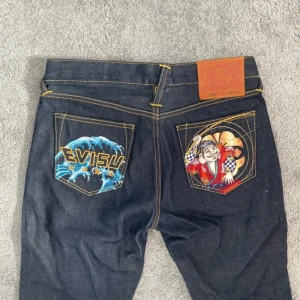 Evisu jeans med broderade bakfickor - Mörkblå jeans från Evisu med unika broderier på bakfickorna – ena med våg och logga, andra med japansk figur. Klassisk femficksmodell, kontrastsömmar och läderpatch i midjan. Riktigt snygga jeans för dig som vill sticka ut.