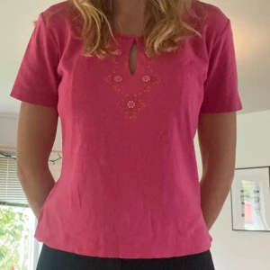 Rosa topp med broderi från KappAhl - Rosa topp från KappAhl (FRIDAY) med kort ärm och broderade blommor i orange och rosa vid halsringningen. Rund hals med liten öppning framtill.