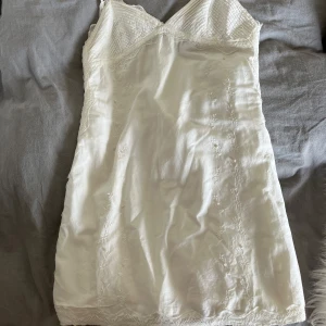 Vit kort klänning med spetsdetaljer från Zara  - Supersöt vit kort klänning med tunna axelband och v-ringning. Klänningen har fina spetsdetaljer upptill och längs nederkanten samt diskret broderade blommor framtill. Perfekt för sommarens varma dagar eller studentfirande.