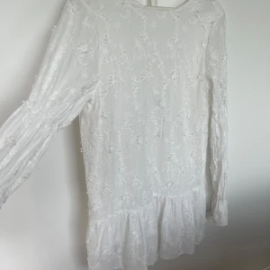 Vit kortklänning med volang - Superfin vit kortklänning med broderade blommor, långa ärmar och volang nertill. Klänningen har djup rygg med knytband och är gjord i ett lätt tyg som känns luftigt. Perfekt för sommarens alla tillfällen!