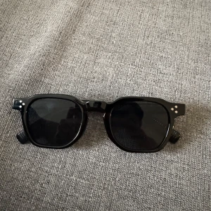 Svarta solglasögon - Snygga svarta solglasögon med fyrkantiga bågar och coola silverdetaljer på skalmarna. Bågarna har tre små silvriga prickar i hörnen och ett mönstrat inslag på insidan av skalmarna. Perfekta för att lyfta din stil i sommar.