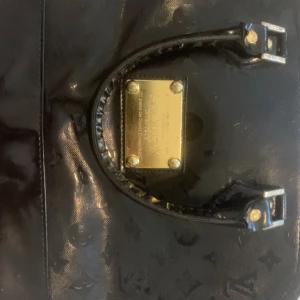 Svart handväska i skinn - Säljer en svart handväska i glansigt skinn med klassiskt monogrammönster och guldfärgade metalldetaljer. Väskan har två handtag med spännen och röd- och svartmönstrat tygfoder invändigt. Perfekt för dig som vill ha en ikonisk och lyxig accessoar.