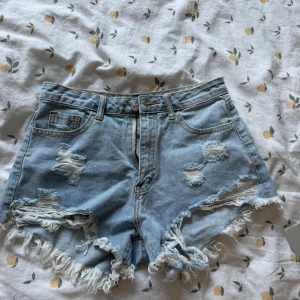 Ljusblå slitna jeansshorts med fransar - Snygga ljusblå jeansshorts med hög midja och slitna detaljer framtill. Råa fransar längs bensluten ger en cool vibe. Klassisk femficksmodell med dragkedja och knapp. Perfekta för sommardagar när du vill ha en avslappnad och trendig look.