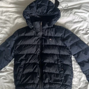 Mörkblå pufferjacka från GANT - Snygg mörkblå pufferjacka från GANT med huva och dragkedja framtill. Jackan har två fickor med knappstängning och GANT-logga på bröstet. 