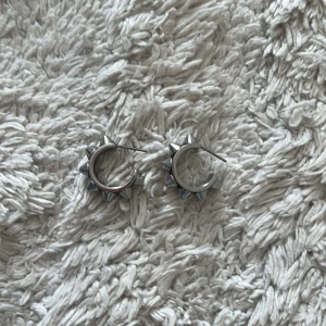 Silvriga örhängen med nitar - Coola silvriga örhängen i metall med rund form och flera spetsiga nitar runt hela ringen. Ger en edgy vibe och passar perfekt om du gillar alternativ stil eller vill sticka ut lite extra.