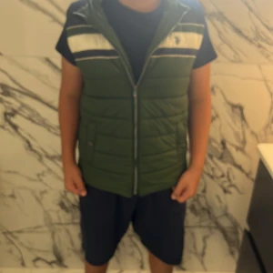 Grön dunväst från U.S. Polo Assn. - Snygg grön dunväst från U.S. Polo Assn. med marinblå och vita ränder upptill, dragkedja framtill och avtagbar huva. Västen har broderad logga på bröstet och två fickor med knappstängning. Perfekt för lager-på-lager och chill stil.