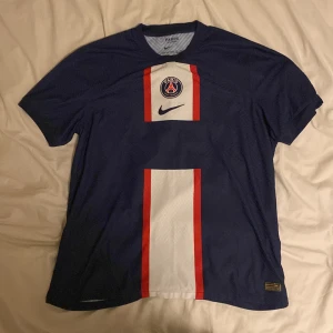 PSG Nike fotbollströja marinblå - Snygg Paris Saint-Germain fotbollströja från Nike i marinblått med vit och röd vertikal panel på framsidan. PSG-logga och Nike swoosh på bröstet. Tillverkad i lätt och ventilerande material, perfekt för match eller träning.