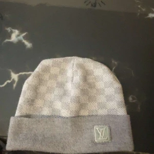 Grå rutig mössa från Louis Vuitton - Snygg grå mössa från Louis Vuitton med klassiskt rutig mönster och vikt kant. Mössan har en diskret LV-logga framtill och är tillverkad i mjukt stickat material som känns lyxigt och bekvämt. Perfekt accessoar för att lyfta din vinterstil.