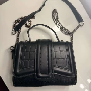Svart axelväska med kedja och krokoprägling - Snygg svart axelväska med krokopräglat mönster och silverfärgad kedjerem. Väskan har ett top handtag och ett stort lock med magnetknäppning.