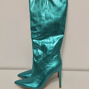 Turkosa höga stövlar med klack - Statementstövlar i metallic turkos med ormskinnsmönster och spetsig tå. Höga skaft och smal hög klack ger en riktigt edgy vibe. Perfekta för dig som vill sticka ut och addera färg till outfiten.