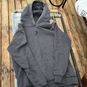 Grå hoodie från Peak Performance - Säljer en stilren grå hoodie från Peak Performance med stor logga på ryggen och mindre logga på bröstet. Tröjan har huva, känguruficka och ribbade muddar. Perfekt för chill dagar eller när du vill ha en clean look.