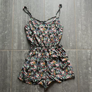 Blommig linnetopp från H&M Divided - Supersöt blommig body från H&M Divided med tunna axelband och elastisk midja. Svart botten med färgglada blommor i rosa, blått, grönt och orange. 