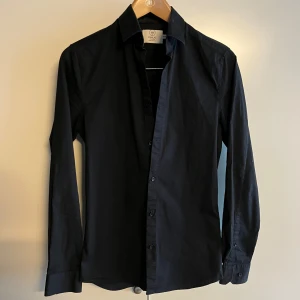 Emilio skjorta svart - Klassisk svart skjorta från Emilio i storlek XS. Skjortan har lång ärm, knappar framtill och en stilren krage. Tillverkad i bomull för en skön känsla och snygg passform. Perfekt för dig som gillar en enkel och clean stil.