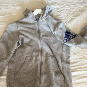 Grå hoodie från Adidas med blå logga - Snygg grå hoodie från Adidas med dragkedja och huva. Stor blå Adidas-logga på ena ärmen ger en sportig vibe. Mjuk insida och två fickor framtill. Perfekt för chill eller träning.