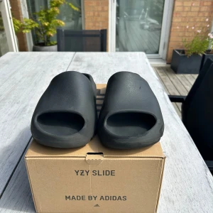 Adidas Yeezy Slide Onyx strl 46 - Svarta Adidas Yeezy Slide Onyx i storlek 47. Slidesen har en chunky, minimalistisk design med öppen tå och räfflad sula för extra grepp. Tillverkade i syntetmaterial som ger en clean och modern vibe. Perfekta för chill dagar eller när du vill ha en stilren look.