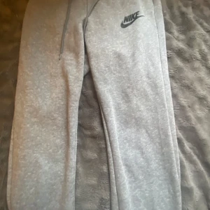 Grå mjukisbyxor från Nike - Snygga grå mjukisbyxor från Nike med klassisk logga på vänster lår. Byxorna har dragsko i midjan för perfekt passform och är tillverkade i mjukt bomullsmaterial. Perfekta för chill eller träning, med en clean och sportig look.