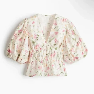 Blommig blus med puffärm och v-ringning - Supersöt blommig blus i vitt med rosa och gröna detaljer. Blusen har v-ringning, smockad midja och puffiga trekvartsärmar som ger en romantisk vibe. Perfekt för dig som gillar en feminin och trendig look.