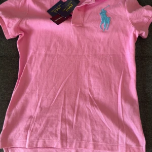Rosa pikétröja från Ralph Lauren M - Snygg rosa pikétröja från Ralph Lauren i modellen The Skinny Polo. Tröjan har klassisk krage, korta ärmar och en stor turkos broderad logga på bröstet. Tillverkad i bomull för en skön och luftig känsla. Perfekt för dig som gillar en sportig och fräsch stil.