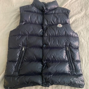 Mörkblå dunväst från Moncler - Snygg mörkblå dunväst från Moncler med quiltad design och hög krage. Västen har två dragkedjefickor framtill och klassisk Moncler-logga på bröstet. Perfekt för lager-på-lager och riktigt street. Materialet är glansigt och känns lyxigt.