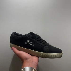 Svarta Saint Laurent sneakers strl 44 - Svarta sneakers från Saint Laurent med broderad logga på sidan och guldfärgad text på plösen. Skorna har svarta snören, vit platt sula och är tillverkade i en mix av skinn och textil. Klassisk och stilren modell som passar till det mesta. Använda 2-3 gånger och säljer dem för att dem inte satt bra på mig. PRIS GÅR ATT DISKUTERAS!!!