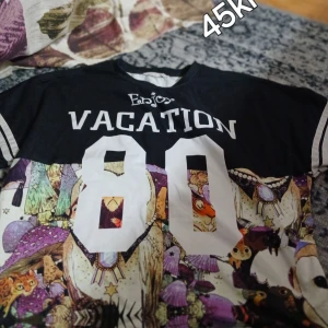 Vacation 80 t-shirt med djurmönster - Cool t-shirt från Enjoy med svart överdel, stora vita tryck med texten VACATION och siffran 80. Nedre delen har ett färgglatt djur- och naturmönster i lila, beige, grön och orange. Klassisk rund hals och korta ärmar med vita ränder.